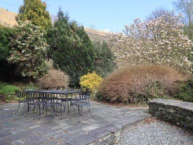 An outdoor patio with a black metal table and eight chairs surrounded by various trees and bushes at Rawfell in Great Langdale