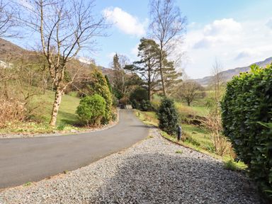 An asphalt driveway with a gravel shoulder lined with bushes and trees in a rural landscape at Rawfell in Great Langdale