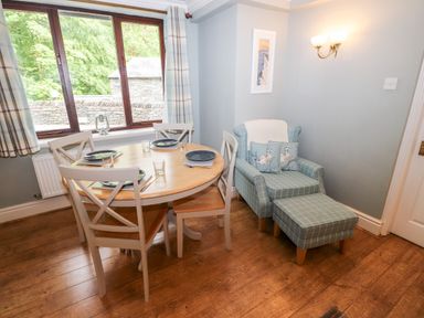 A dining area with a round wooden table set for four and an armchair with an ottoman in a room at Babbling Brook in Ambleside