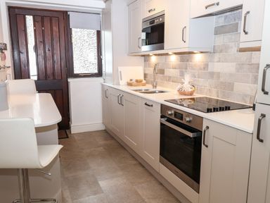 A kitchen with white cabinets a built in oven and microwave a sink and a white dining table with chairs and a dark wooden door at Babbling Brook in Ambleside