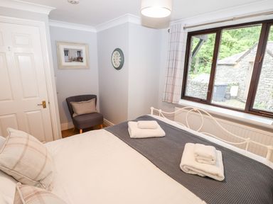 A bedroom with a bed, chair, wall clock, framed picture, window with curtains, and folded towels at Babbling Brook in Ambleside