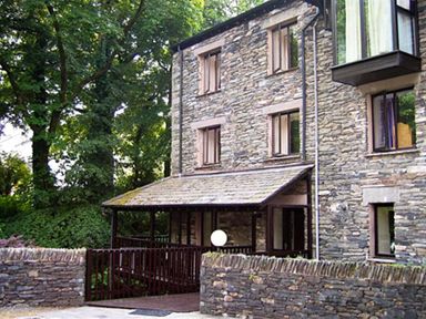 Stone house with multiple windows and a covered porch behind a stone wall and gate surrounded by trees at Over Ghyll Ambleside