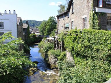 A narrow river flowing between stone buildings with greenery and a water wheel in Ambleside