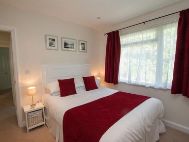 A bedroom with a double bed with red cushions and a red throw a bedside table with a lamp and a window with red curtains at Bield How in Ambleside