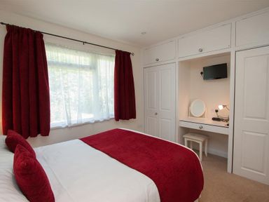 A bedroom with a bed with red pillows and blanket next to window with red curtains and built-in wardrobe with vanity and TV at Bield How in Ambleside