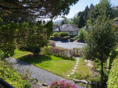 A garden with grass, shrubs, trees, a gravel path, and stone slabs leading to a road with houses beyond at Bield How in Ambleside