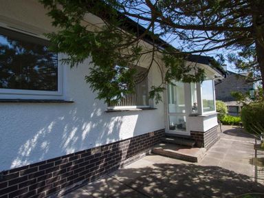 Outside view of a house with white walls and dark brick base under trees and a paved walkway at Bield How in Ambleside