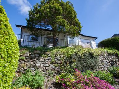 A stone retaining wall with plants and flowers in front of a white house with a tree and metal railing at Bield How in Ambleside