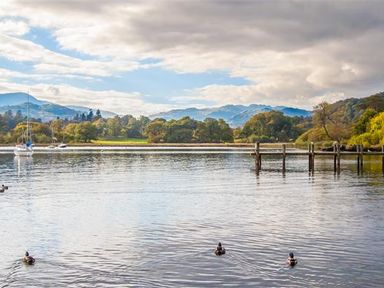 A lake with ducks sailing boats a wooden jetty and trees with mountains in the background at Bield How in Ambleside
