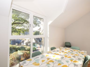 A dining area with a table covered in a leaf patterned cloth and green chairs next to a large window showing houses and trees at Garden House in Ambleside