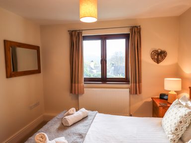 A bedroom with a window with curtains a bed with rolled towels and pillows a wooden nightstand with a lamp and a heart decoration on the wall at Garden House in Ambleside