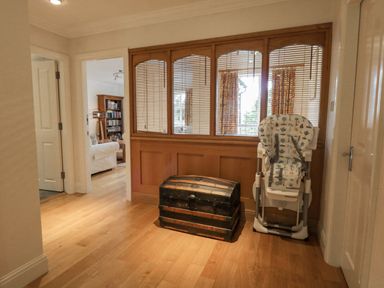 A hallway with wooden floor a black chest and a folded baby high chair by wooden window panels looking into a sitting room at Orchard House in Ambleside
