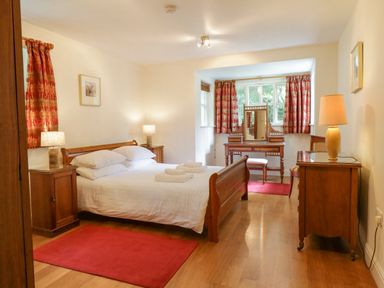 A bedroom with a wooden bed and white bedding two bedside tables with lamps a dressing table with a mirror and a chair red curtains and rugs at Orchard House in Ambleside