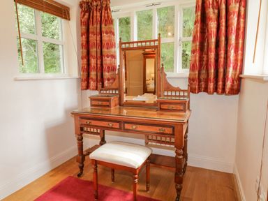 A wooden dressing table with mirror and stool in a corner with red patterned curtains at Orchard House Ambleside