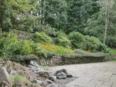 A paved patio area with a rock garden and stone wall with shrubs and trees at Orchard House in Ambleside