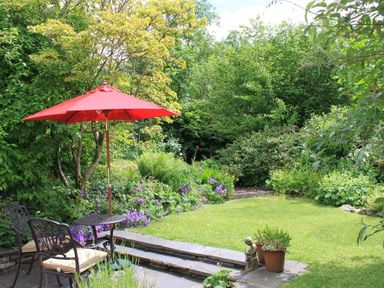 A garden with a red patio umbrella over a metal table and chairs surrounded by green plants and flowers at Bank Foot in Ambleside
