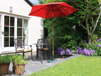A patio with two chairs a table and a red umbrella next to a white house with glass doors and garden plants at Bank Foot Ambleside