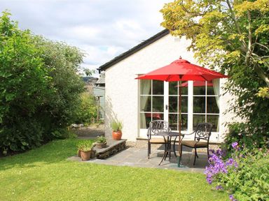 A garden patio with a round table and two chairs under a red umbrella by a white house with glass doors at Bank Foot in Ambleside