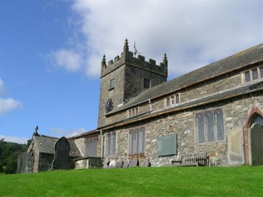 A stone church building with a clock tower grave markers and a bench on a grassy hill at Janes Cottage in Hawkshead