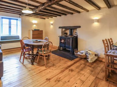 A dining area with a round wooden table, chairs, a wood stove, a chest of drawers, and a window with a bench at Janes Cottage in Hawkshead