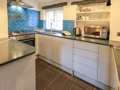 A kitchen with blue tiled backsplash a gas stove oven microwave kettle and open shelves with glassware at Janes Cottage in Hawkshead