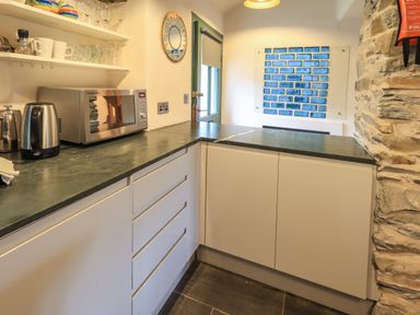 A kitchen with white cabinets a microwave kettle glassware on shelves and a blue tiled artwork on the wall at Janes Cottage in Hawkshead