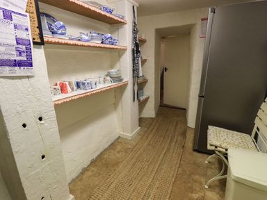 A kitchen with open shelves holding dishes and mugs a refrigerator a bench with a cushion and a rug on the floor at Nurses Cottage in Hawkshead