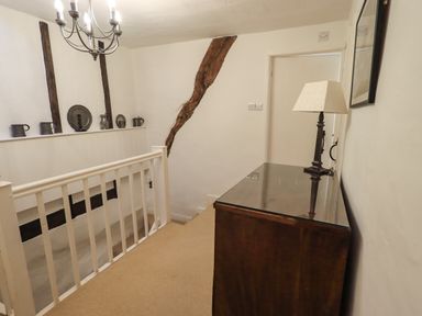 A hallway with a wooden railing, a chest of drawers with a lamp, and exposed wooden beams at Nurses Cottage in Hawkshead