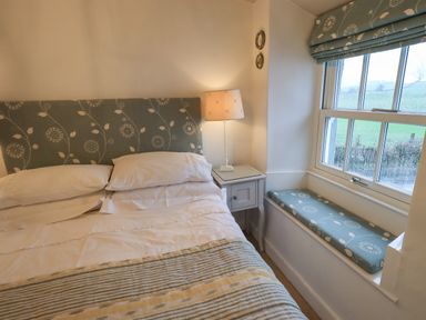 A bedroom with a bed having patterned headboard and striped bedspread a nightstand with lamp and a window seat at Nurses Cottage in Hawkshead