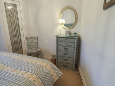 A bedroom corner with a striped bedspread a grey chest of drawers a round mirror a lamp a chair and a door at Nurses Cottage in Hawkshead