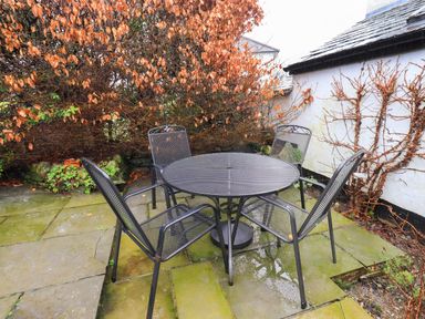 An outdoor patio with a round metal table and four matching chairs surrounded by dried foliage at Nurses Cottage in Hawkshead