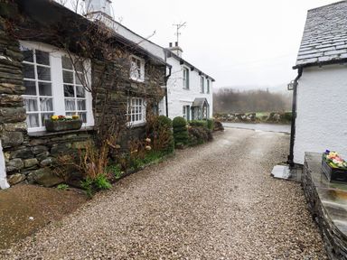 A gravel driveway between two stone and white plaster houses with flowers in window boxes at Nurses Cottage in Hawkshead