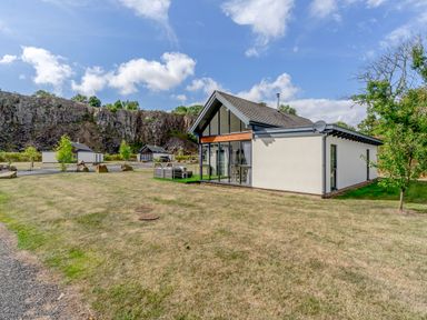 A white house with a slanted roof and large windows on a grassy plot with rock cliffs in the background at Wood Sage in Bamburgh