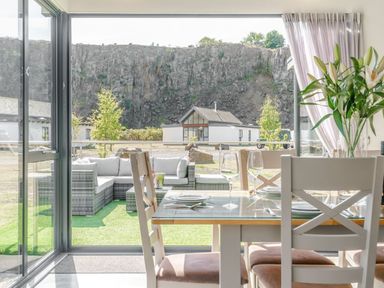A dining area with a glass table and chairs next to a vase of lilies and a glass door opening to an outdoor seating area with a rocky cliff and small buildings in the background at Wood Sage in Bamburgh