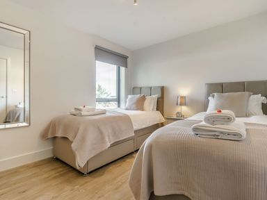 A bedroom with two single beds with beige bedspreads and pillows a bedside table with a lamp and an alarm clock and a wall mirror at Wood Sage in Bamburgh
