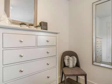 A white dresser with small potted plants and a box on top next to a gray chair with a heart-shaped pillow and a wall mirror at Wood Sage in Bamburgh