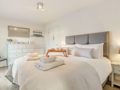A bedroom with a gray headboard bed with white linens and towels on it a wooden tray with breakfast on the bed a white dresser with a mirror above it and a chair in the corner at Wood Sage in Bamburgh