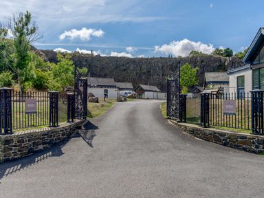 The entrance gate to a lodge complex with a driveway leading to several buildings and rocky cliffs in the background at Brada Lodges Bamburgh