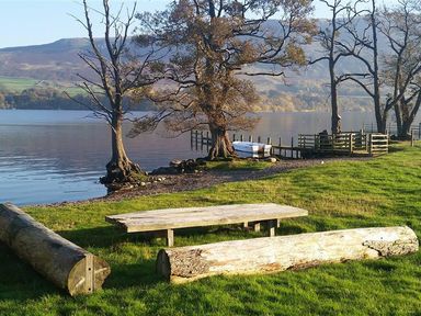 A picnic table with log benches by a lake with trees and a small dock at Low House in Ullswater