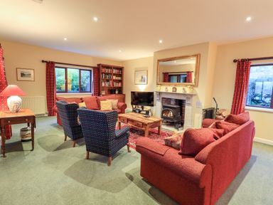 A living room with red sofas, two patterned armchairs, a wooden coffee table, a fireplace, a TV, a bookcase, and windows with red curtains at Low House in Ullswater