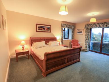 A bedroom with a wooden bed two bedside tables with lamps an armchair and large windows with floral curtains at Low House Ullswater