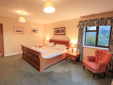 A bedroom with a wooden bed frame two bedside tables with lamps a pink armchair and a window with patterned curtains at Low House Ullswater