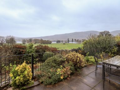 A patio with a table and chairs overlooking a garden with shrubs and a view of fields and distant hills at Low House in Ullswater