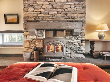A wood burning stove set in a stone fireplace with a red ottoman and an open book in the foreground at High Lindeth in Bowness