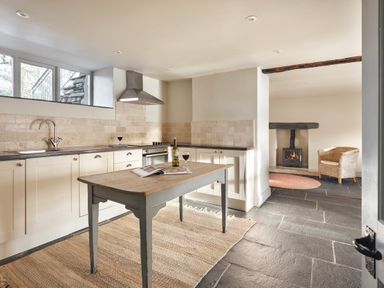 A kitchen with a wooden table a bottle and glasses of wine and a living area with a chair and fireplace at High Lindeth in Bowness