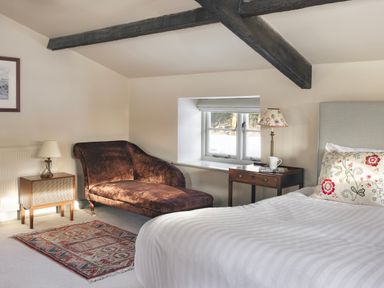 A bedroom with a chaise lounge armchair a bed with floral cushions and wooden side tables at High Lindeth in Bowness