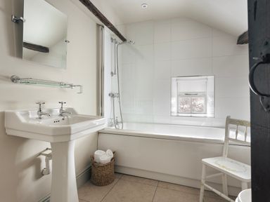 A bathroom with a pedestal sink, bathtub with shower, small window, basket with towels, and wooden chair at High Lindeth in Bowness