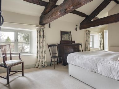 A bedroom with wooden beams two windows floral curtains a bed two chairs and a dark wooden dresser with a mirror at High Lindeth in Bowness