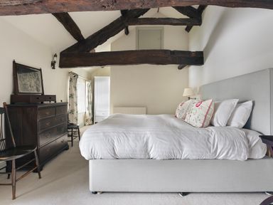 A bedroom with a large bed wooden beams on the ceiling a dark wood dresser two chairs a white wardrobe and a window at High Lindeth in Bowness