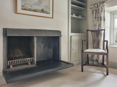 A fireplace next to a wooden chair with floral curtains by a window at High Lindeth in Bowness